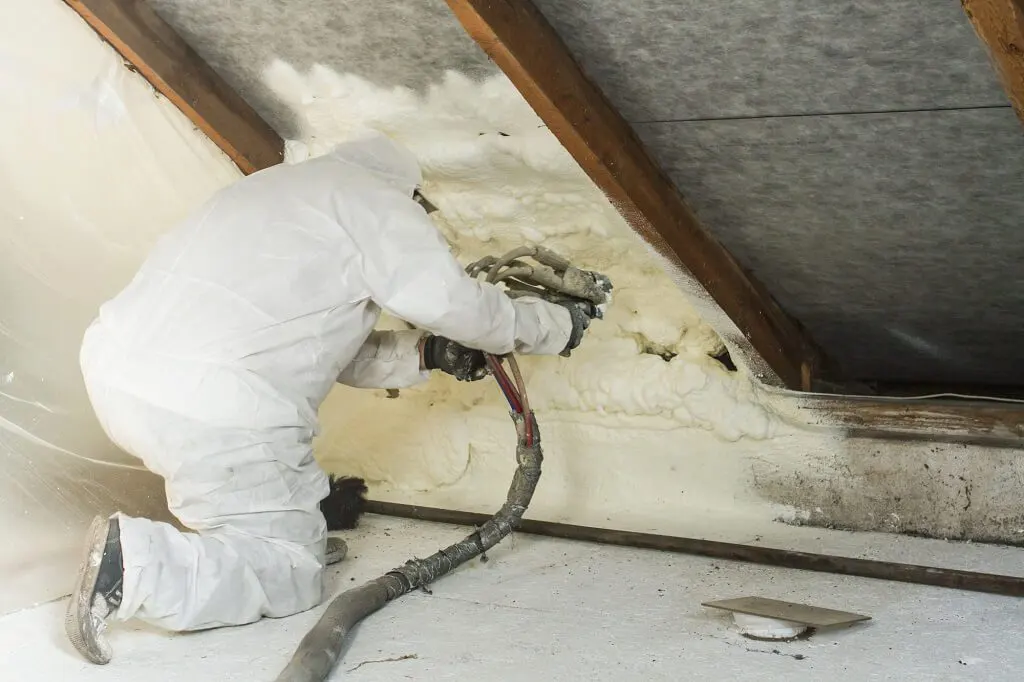 Technician in a white protective suit spraying foam insulation onto an attic roof