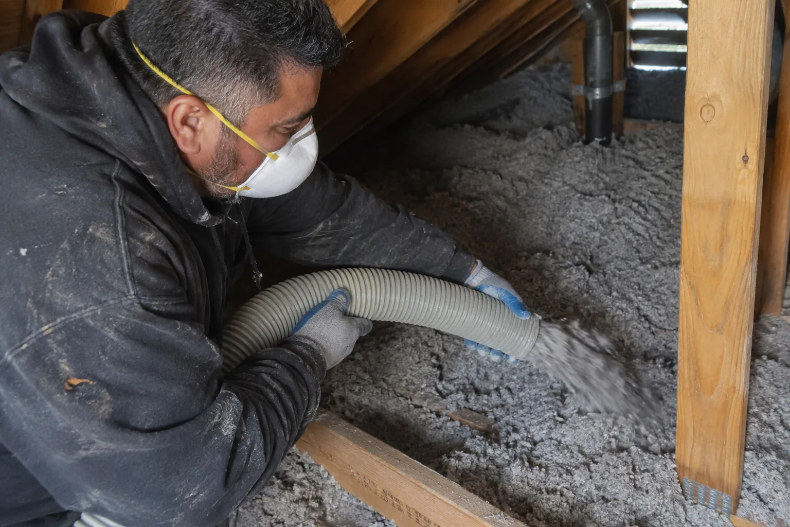 Blowing cellulose insulation across attic floor joists near a vent stack, building up even coverage with a hose.