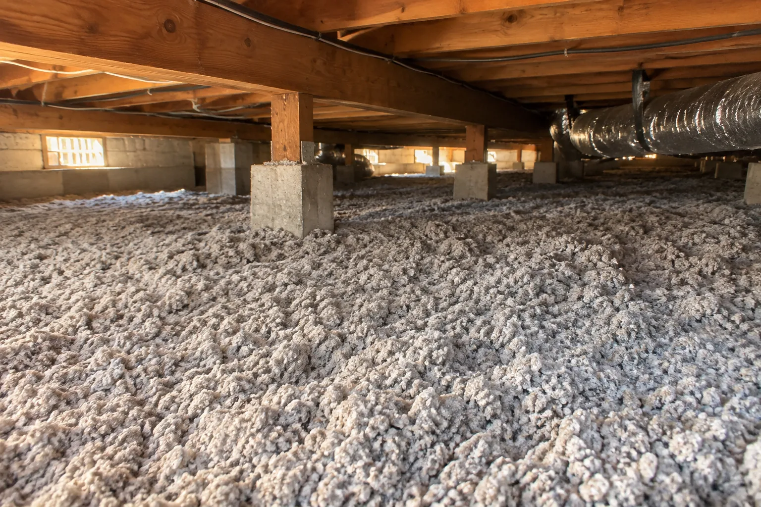 A crawl space floor covered with a thick, even layer of cellulose insulation between the joists and support piers.