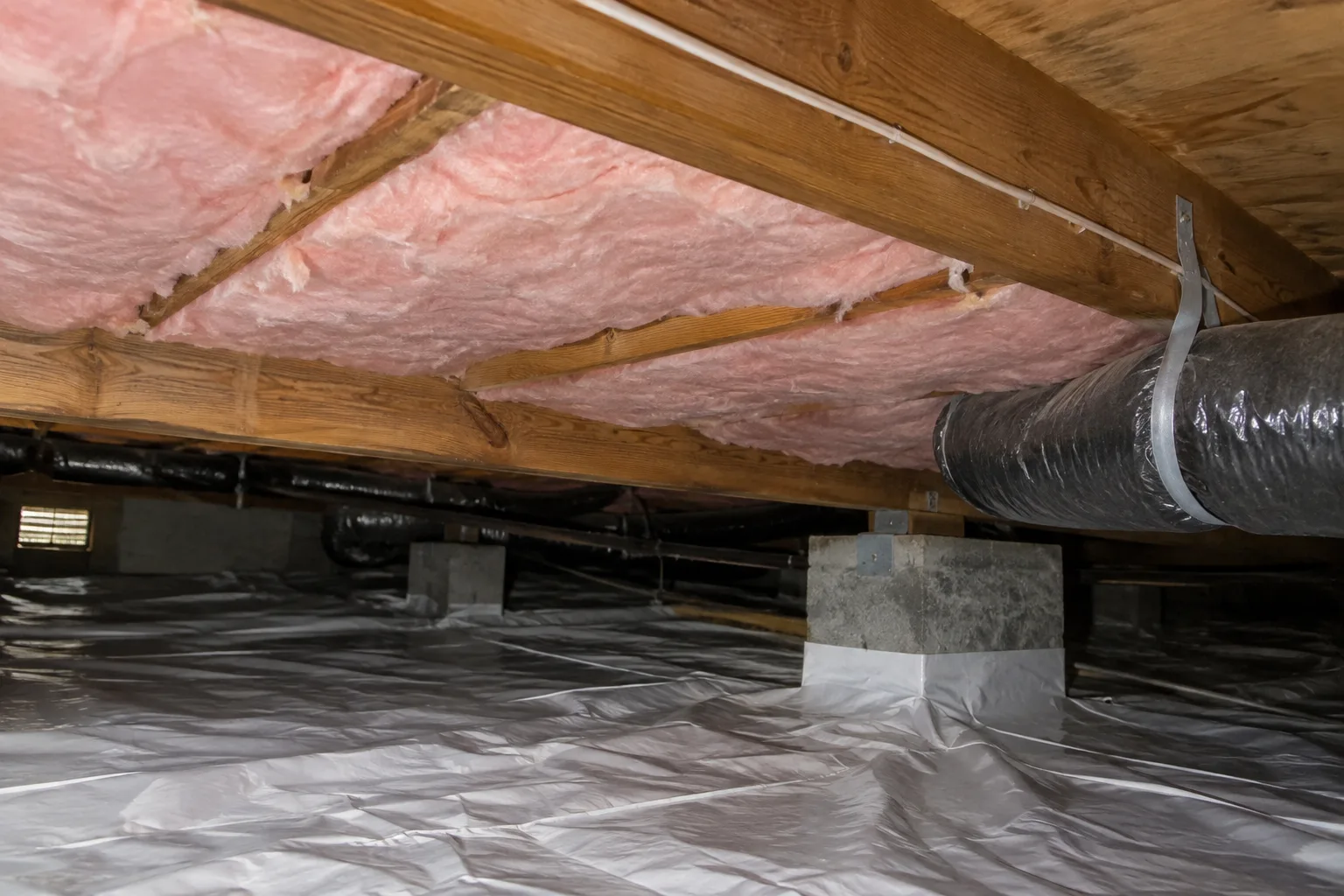 New fiberglass batt insulation installed between crawl space floor joists above a sealed vapor barrier and insulated ductwork.