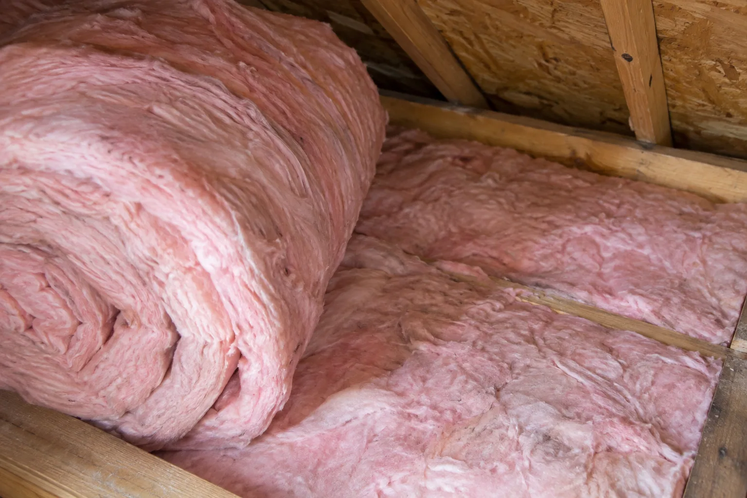 Close-up of a fiberglass insulation roll laid between attic joists, showing full-depth coverage across the attic floor.